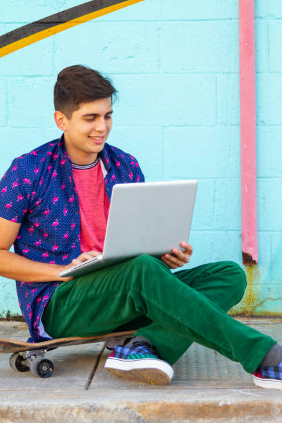 Portrait of young latin college university happy handsome male student studying abroad and gap year enjoying his time in the United States reading an article in his laptop using 5G technology while taking a look around Wynwood District Miami Design District, Miami neighborhood in his skate.
He's wearing a colorful outfit: Tropical shirt, pink t-shirt and green jeans.
Education concept. Studying Abroad and Gap Year. Copy Space. retrato de un joven universitario latino feliz y guapo estudiante universitario que estudia en el extranjero y un año sabático disfrutando de su tiempo leyendo un artículo en su tableta digital utilizando la tecnología 5g mientras echa un vistazo al di