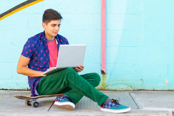 Portrait of young latin college university happy handsome male student studying abroad and gap year enjoying his time in the United States reading an article in his laptop using 5G technology while taking a look around Wynwood District Miami Design District, Miami neighborhood in his skate.
He's wearing a colorful outfit: Tropical shirt, pink t-shirt and green jeans.
Education concept. Studying Abroad and Gap Year. Copy Space. retrato de un joven universitario latino feliz y guapo estudiante universitario que estudia en el extranjero y un año sabático disfrutando de su tiempo leyendo un artículo en su tableta digital utilizando la tecnología 5g mientras echa un vistazo al di