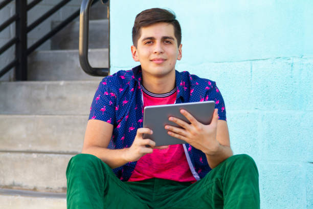 Portrait of young latin college university happy handsome male student studying abroad and gap year enjoying his time in the United States reading an article in his digital tablet using 5G technology while taking a look around Wynwood District Miami Design District, Miami neighborhood in his skate.
He's wearing a colorful outfit: Tropical shirt, pink t-shirt and green jeans.
Education concept. Studying Abroad and Gap Year. Copy Space. retrato de un joven universitario latino feliz y guapo estudiante universitario que estudia en el extranjero y un año sabático disfrutando de su tiempo leyendo un artículo en su tableta digital utilizando la tecnología 5g mientras echa un vistazo al di