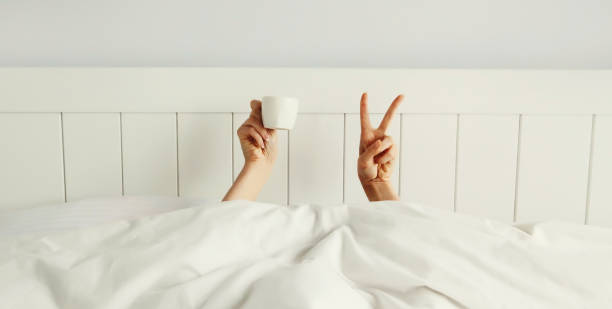 istock Cheerful lazy woman waking up after sleeping lying in bed holding cup in her hands asks for coffee 2159023883