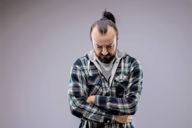 Studio portrait of a bearded man with arms crossed looking down, expressing emotions of sadness, worry and contemplation