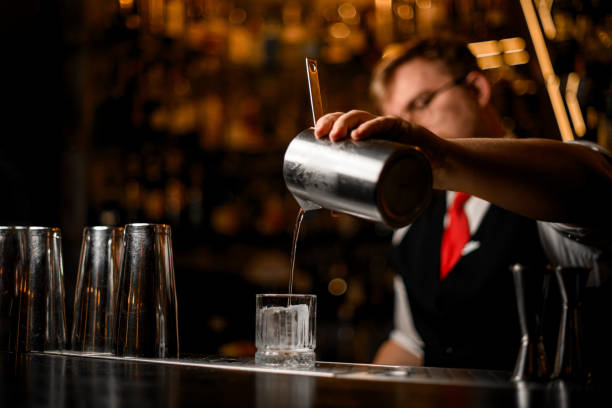 istock Selective focus on glass tumbler with ice cube into which bartender pours clear cocktail 2159059042