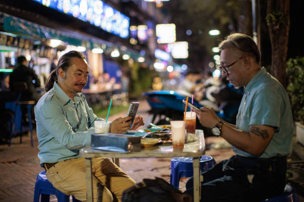 Male friends using smart phones after eating street food marketplace while sitting at the table at night in Bangkok, Thailand.