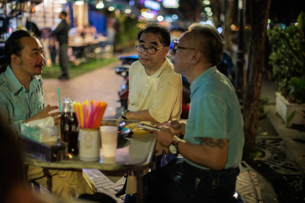 Male friends talking after eating street food marketplace while sitting at the table at night in Bangkok, Thailand.
