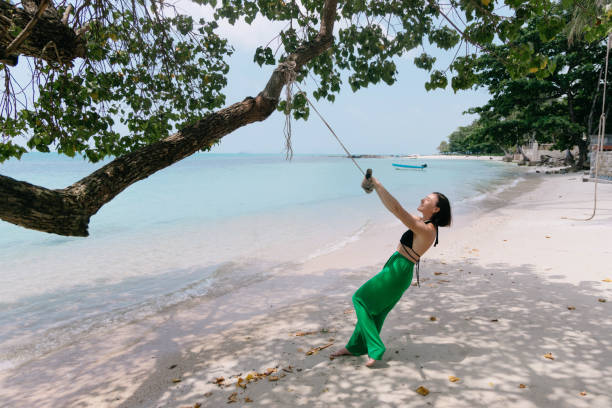 A woman swings on a rope swing that is hanging from a tree on a white sandy beach with turquoise water in the background.