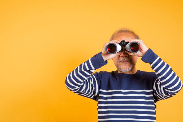 istock Portrait Asian smiling old man looking camera through binoculars studio shot isolated yellow background, senior man pensioner feeling surprised 2159106265