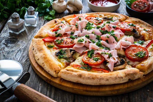 istock Pizza with ham, mozzarella cheese, sliced tomatoes and white mushrooms on wooden table 2159121279