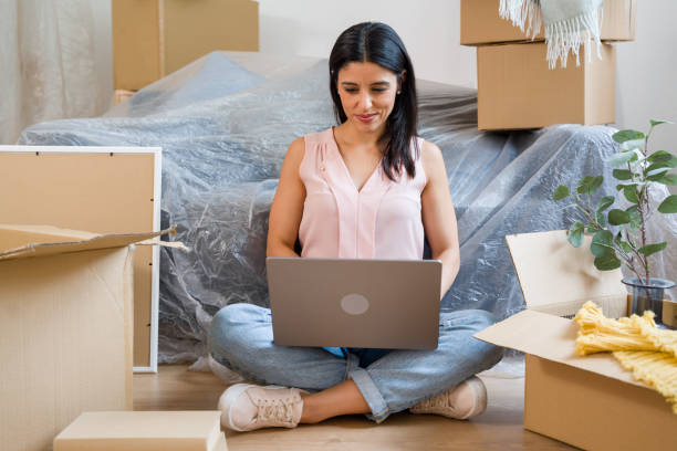 istock Woman using laptop while sitting on floor surrounded by moving boxes 2159125694