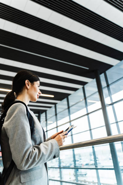 istock Tourist with smartphone, documents and backpack in the airport lounge is looking through the panoramic window. Smiling caucasian woman waiting for flight to start vacation in 2024. 2159129148