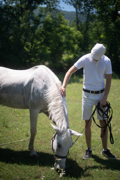 Young man petting white horseback as horse eats grass.