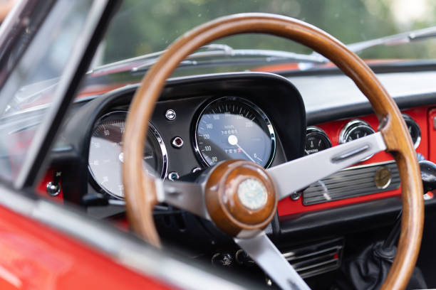 This image showcases the interior of an open vintage car featuring a stylish wooden steering wheel and a traditional red dashboard. The typical round gauges from that era display speed, RPM, and fuel level, evoking a sense of nostalgia and elegance. The retro design and analog instruments make this car a real eye-catcher