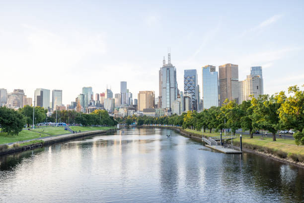 A tranquil river flowing through a modern cityscape during the early evening. The skyline features an array of skyscrapers and urban architecture, with lush green trees lining the riverside, reflecting in the calm water.