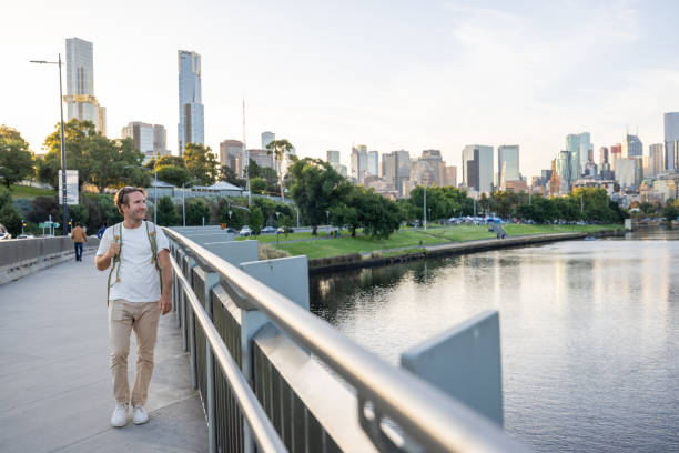 A man with a backpack stands on a bridge overlooking a tranquil river and modern cityscape. The skyline features skyscrapers and urban architecture, with green trees lining the riverside. Perfect for themes of travel, exploration, and urban adventure.