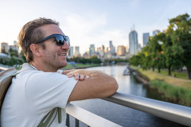 A man wearing sunglasses leans on a bridge railing, smiling as he enjoys the view of a tranquil river and modern cityscape. The skyline features skyscrapers and urban architecture, with greenery lining the riverside.