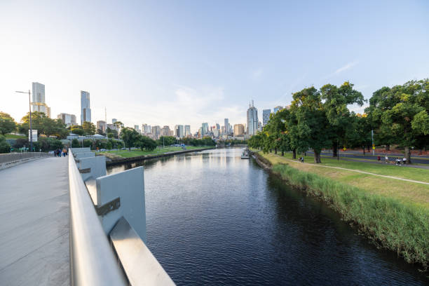 View of a modern city skyline from a bridge overlooking a calm river. The urban landscape is framed by lush greenery and high-rise buildings under a clear sky.