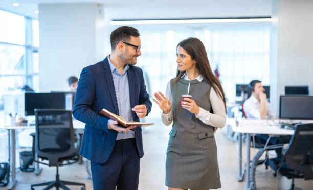 istock Businessman with notebook walking through the office hallway, engaging in conversation with his younger female colleague while sipping on a takeaway coffee. 2159147736