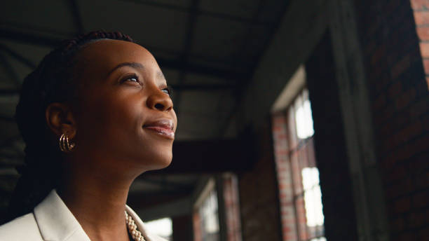 Confident Businesswoman in Modern Industrial Office Environment Portrait of a confident Black woman in a modern industrial office setting, gazing thoughtfully. The image captures feelings of ambition and introspection with brick walls and large windows in the background. portrait power pose stock pictures, royalty-free photos & images