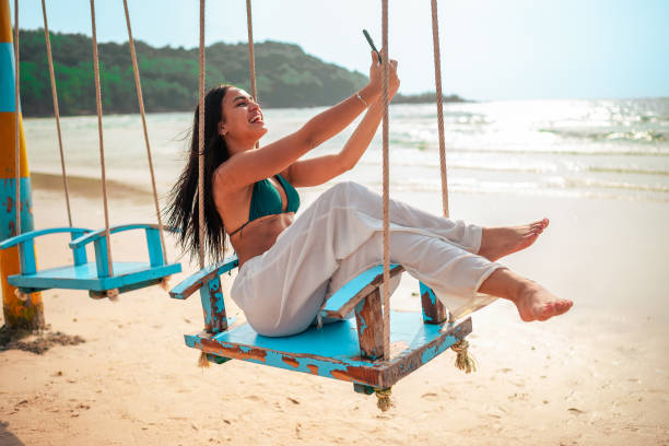 istock Smiling Hispanic Woman on Beach Swing with Smartphone 2159160340
