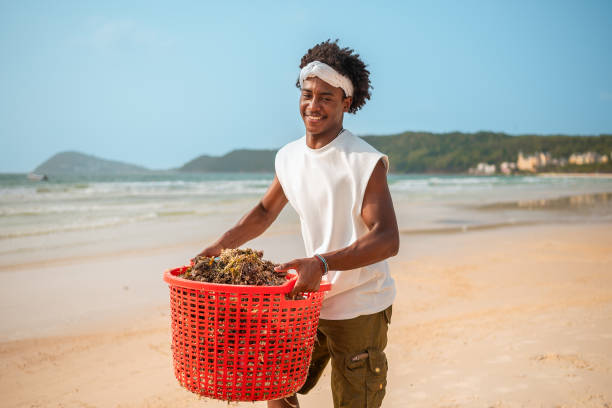 Energetic young adult male of African descent engages in environmental cleanup, cheerfully removing debris from a sunny coastal beach with a red basket in hand.