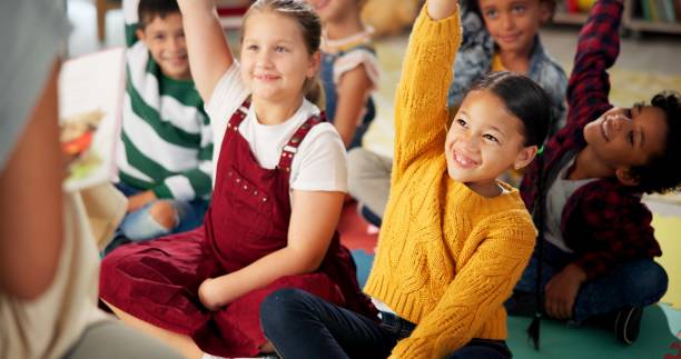 istock Children, teacher and hands up in classroom for reading question or back to school, learning or education. Kids, group and friends on floor for storytelling knowledge or library, growth or studying 2159187486