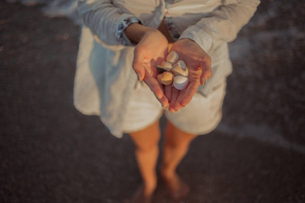 A woman holds shells and pebbles in her palms on the ocean shore.