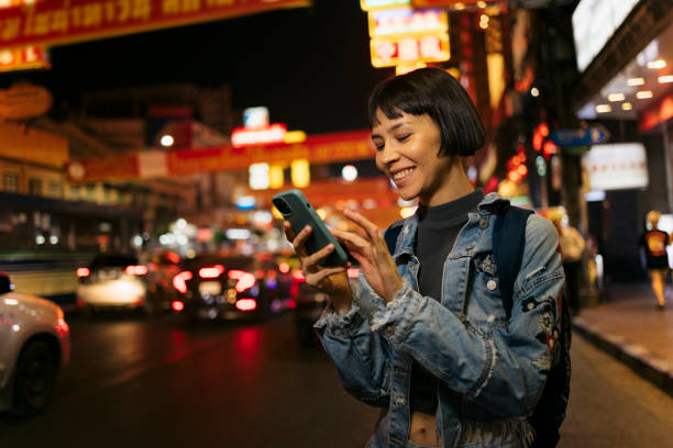 istock Happy young woman using mobile phone while standing on city street at night 2159207720