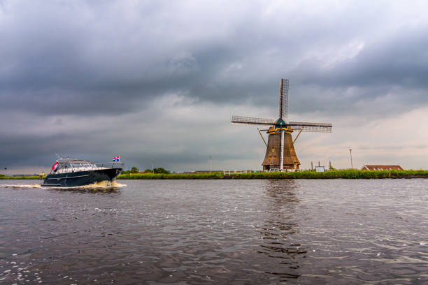Alphen aan den Rijn, Netherlands - May 20 2024: A sloop sails on the Heimanswetring in Alphen aan den Rijn with the Vrouwgeestmolen in the background. A typical Dutch landscape on a cloudy day