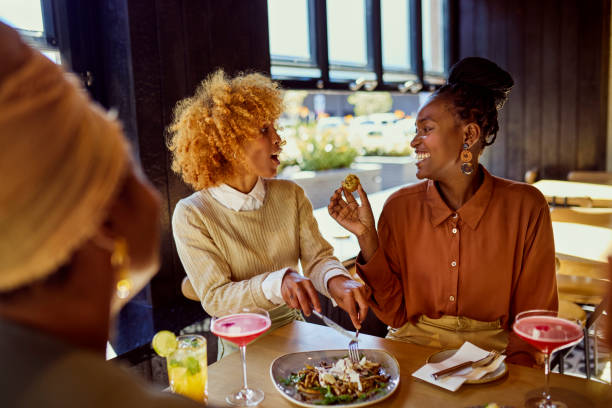 istock Two playful multiracial girlfriends sharing meal for lunch, feeding each other at social gathering in restaurant 2159279364