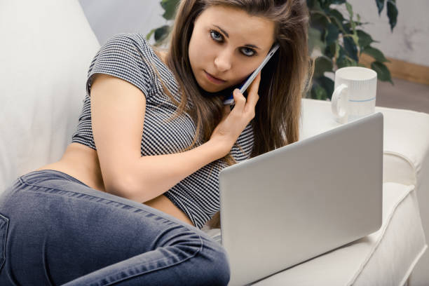 Young woman is lying on her couch, working on her laptop and talking on the phone. She seems to be sad and worried about something
