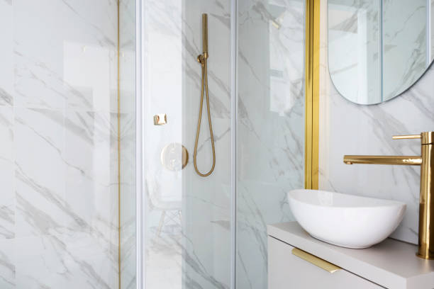 istock Home interior with ceramic bathroom sink on cupboard with gold water tap and shower with tile on the wall with marbled effect.  Interior decor. 2159313313