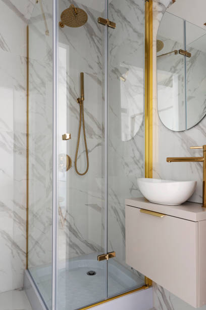 istock Home interior with ceramic bathroom sink on cupboard with gold water tap and shower with tile on the wall with marbled effect.  Interior decor. 2159313317