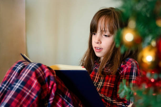 istock Girl reading a book next to Christmas tree 2159313582