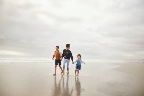 istock Three happy siblings walking and skipping hand in hand on beach 2159317790