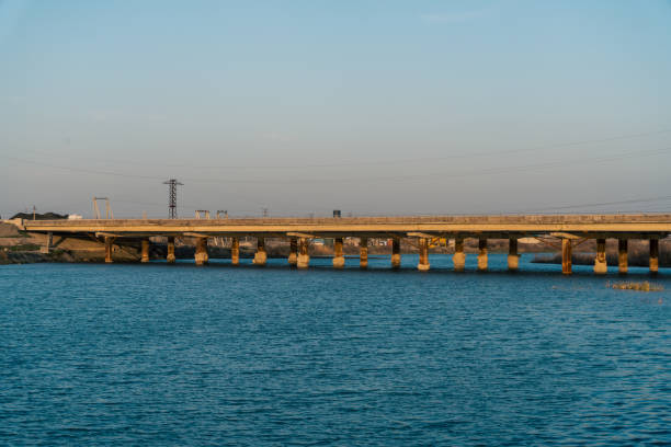 A bridge spans a body of water, with a clear blue sky above. The bridge is long and narrow, with a few pylons and power lines visible in the background. The water is calm and still