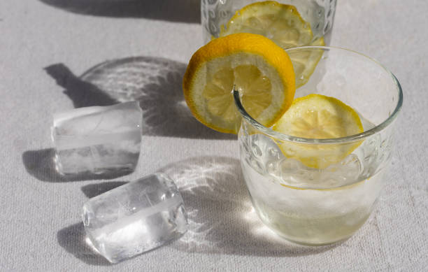 Detail of two glasses filled with cold water and lemon slices, accompanied by ice cubes on a white tablecloth, highlighting freshness and summer refreshment.