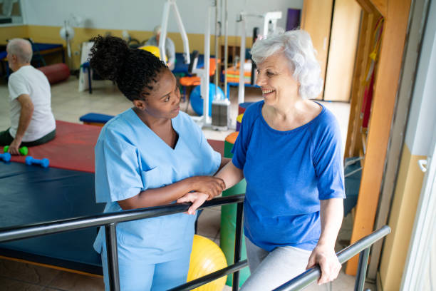 Senior woman guided by therapist in walking exercise between bars at rehab