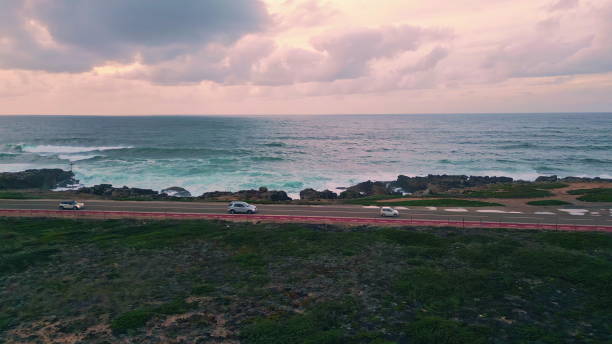 Coastal road stretching beautiful ocean coast washed by stormy turquoise waves drone view. Foamy sea rolling on coast under cloudy sky creating tranquil vivid seascape. Cars riding on asphalt roadway.