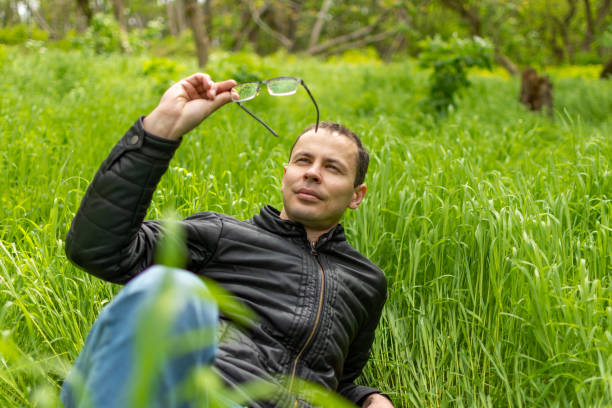 A man lies in the grass with his glasses resting on his head as he enjoys nature. He appears happy surrounded by plants in a natural landscape