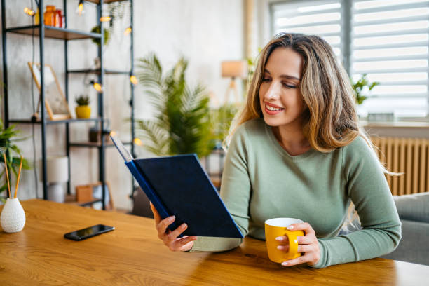 istock Young Woman Reading A Book And Drinking Coffee While Sitting At The Kitchen Table At Home 2159356056