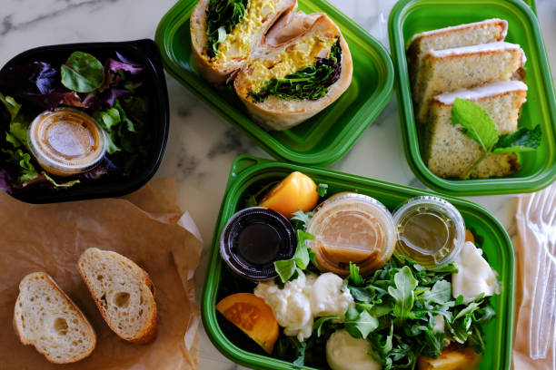 Salads and sandwiches in green and black take out containers on white marble table top. Chicken Curry Sandwich, Tomato Burrata Salad, lemon poppyseed cake, bread and salad