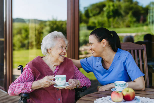 female caregiver spending time with elderly woman, drinking coffee and talking. nurse and senior woman in wheelchair enjoying a warm day outdoors. - ouderenzorg stockfoto's en -beelden
