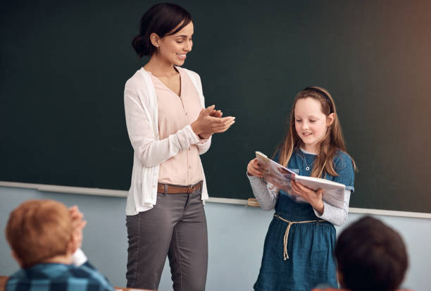 istock Reading, book and girl in classroom, teacher and clapping for student, smile and learning of literature. Proud, woman and applause in school, growth or development of children, knowledge or education 2159398499