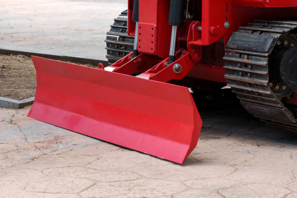 Bulldozer blade of a modern red construction tractor machine with tracks positioned on an urban concrete surface close up