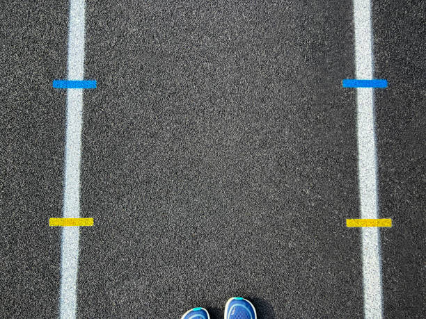 Overhead view of a rubber black running track surface with white lane lines. Yellow and blue dashed lines at the center of view. A running stands at the edge of view. Overhead view of the textured artificial black surface.