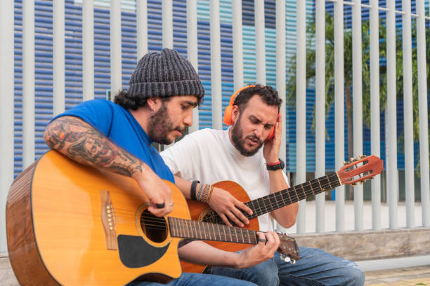 Male teacher with his student sit on stools playing acoustic guitars outside.