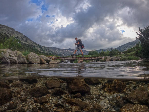 Full length shot of mature man with backpack and hiking poles crossing mountain river over wooden footbridge, shot taken with wearable camera