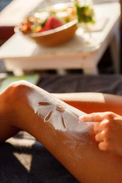 Attractive early 30's smiling blond woman applying suntan lotion onto her skin while sunbathing on the beach. She's wearing bikini,sunglasses and red sun hat, sitting in a deck chair. There are blurry people sunbathing in background.
