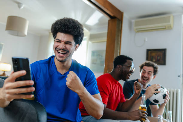 istock Young man using smart phone while watching sports with friends at home 2159477257