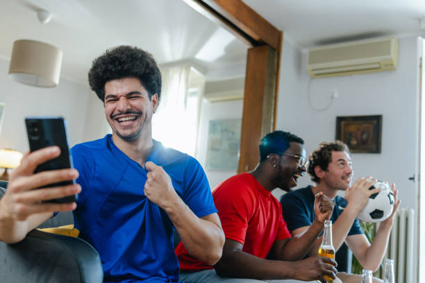 istock Young man using smart phone while watching sports with friends at home 2159477436