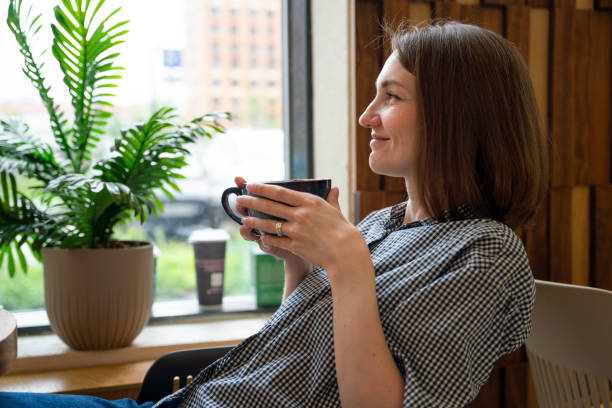 istock Satisfied woman sits with cup coffee at the window in cafe and looking through window 2159488034